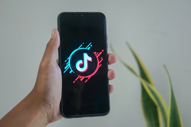 A hand holding a smartphone displaying the TikTok logo on a black screen with a minimalist background.